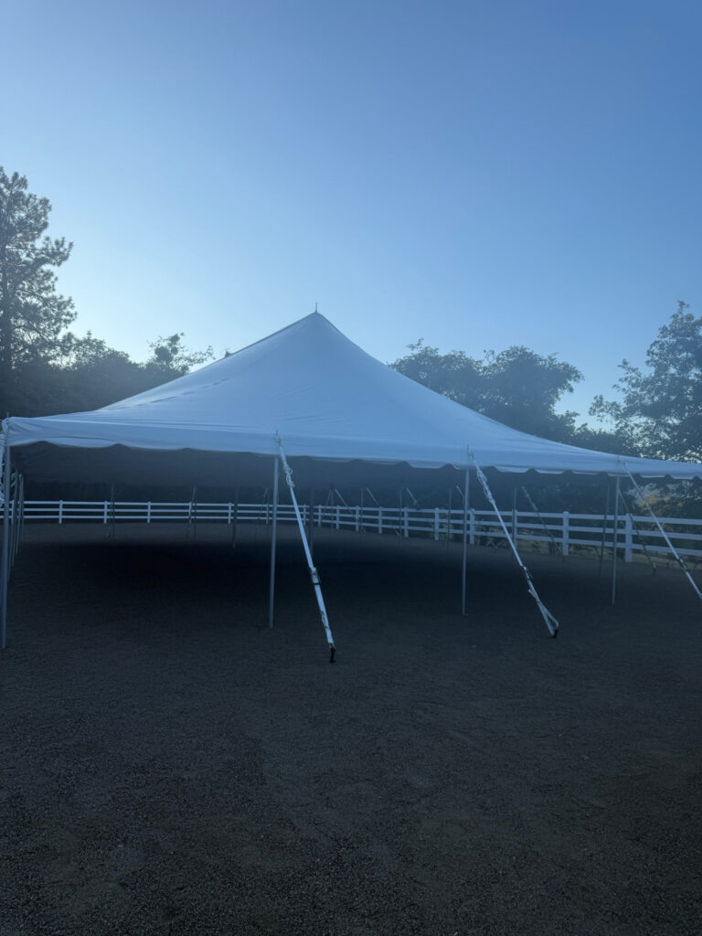 Portrait view of an open-sided white pole tent with perimeter poles, guy lines and a white fence.