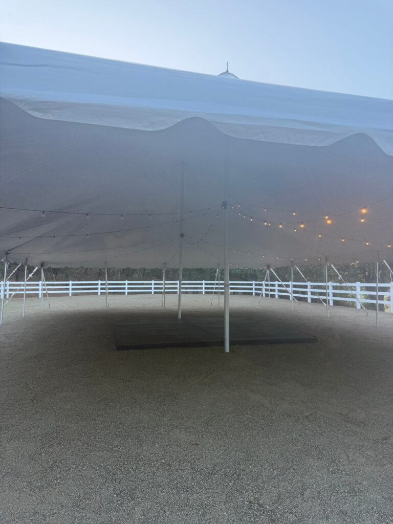 View through a white pole tent opening with illuminated string lights, center poles and a dark rectangular floor area.