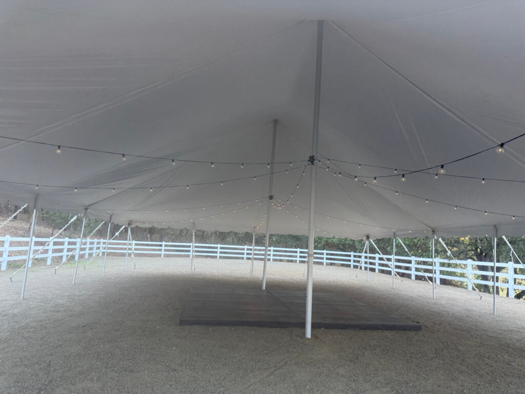 Illuminated string lights beneath a white pole tent, with center poles and a dark rectangular floor area.