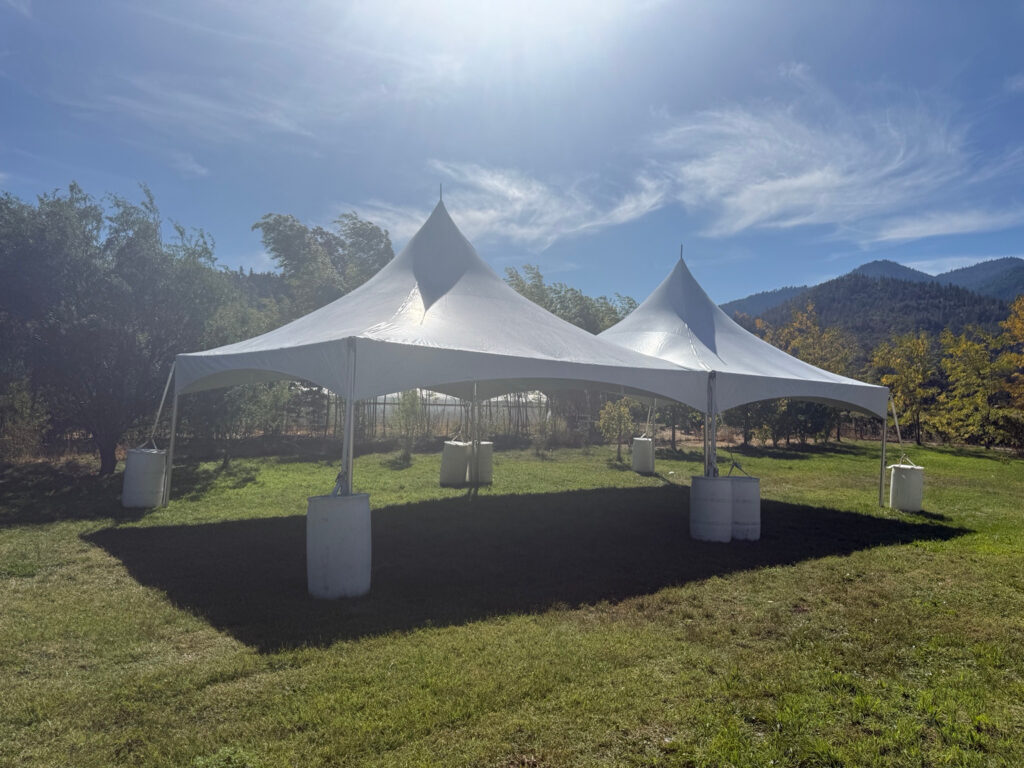 Two white high-peak tents set together on grass, with white barrels beside their support legs.