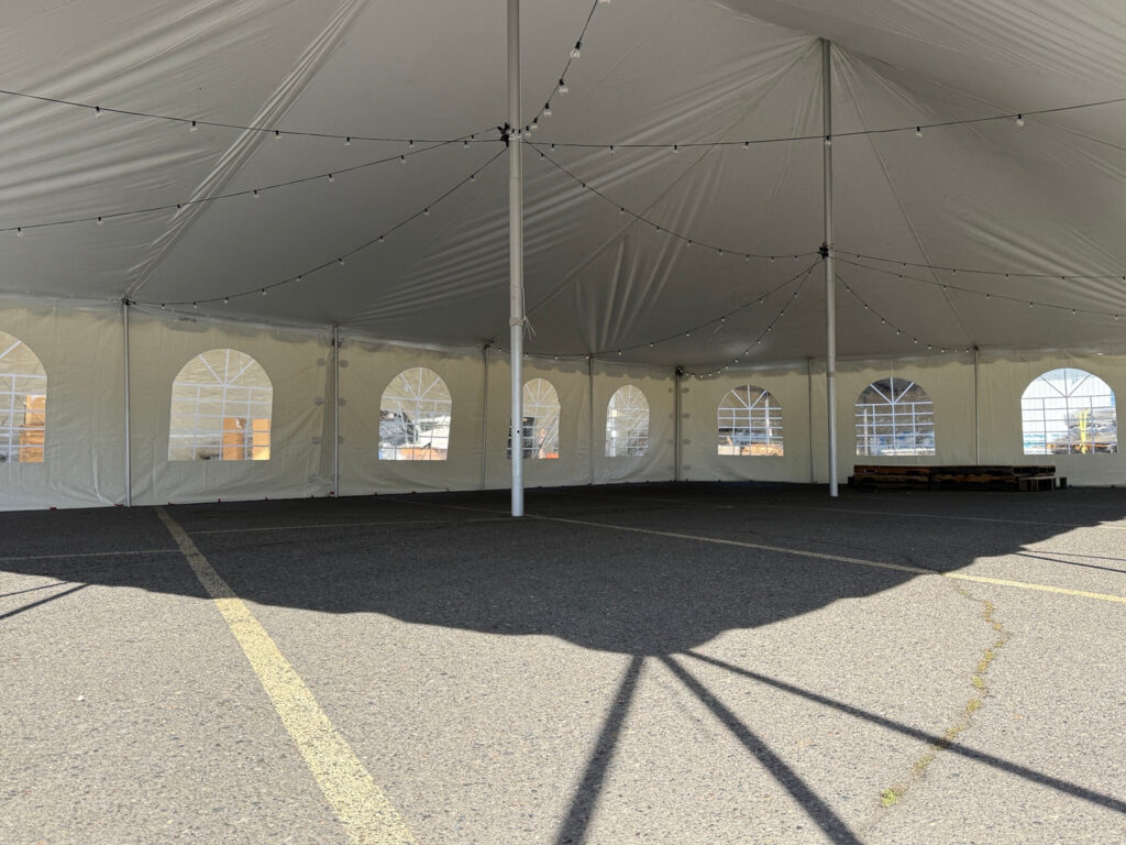Inside a white pole tent with center support poles, arched window sidewalls, string lights and a paved floor.